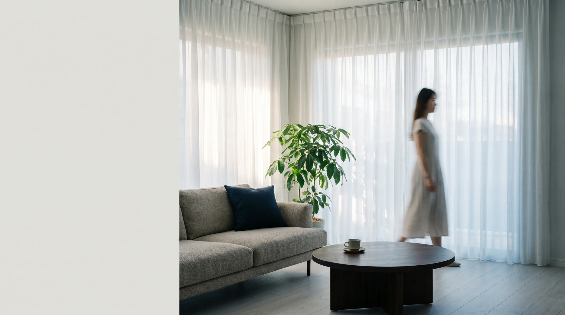 Sala clara com cortinas de voil branco do teto ao chão e uma pessoa passando ao fundo.